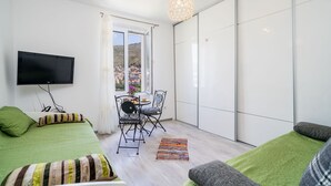Ein modernes Schlafzimmer mit Flachbildfernseher, grüner Bettwäsche, einer Essecke mit Stühlen und Blick auf eine Hügelstadt durch ein großes Fenster.