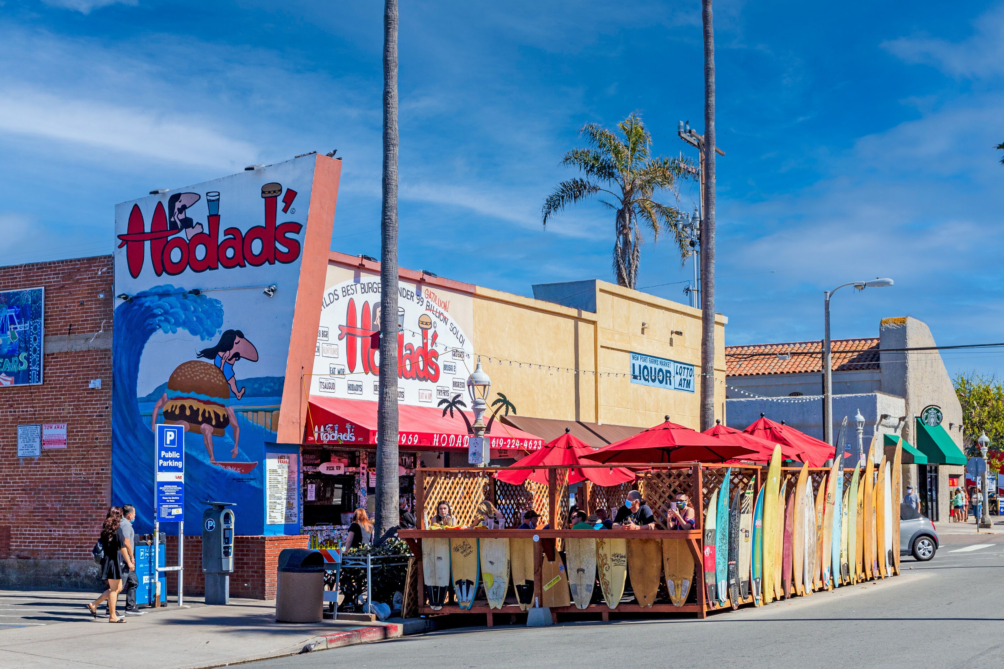 Un restaurant appelé « Hodad’s » dont la façade est ornée d’une fresque représentant un surfeur et une scène de plage. On y voit des palmiers et un ciel bleu clair en arrière-plan.