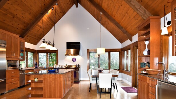 A spacious kitchen with wooden cabinets, a large island, and a dining area with a view of trees. - River's Edge Retreat by North Bend Escapes (Fall City)