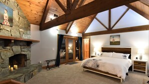 A bedroom with a stone fireplace, wooden beams, a bed with a feathered duvet, and a nightstand with a lamp. - River's Edge Retreat by North Bend Escapes (Fall City)
