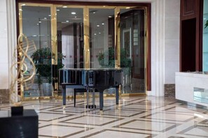 Un piano de cola en un vestíbulo con piso de baldosas geométricas y entrada por puerta de vidrio.