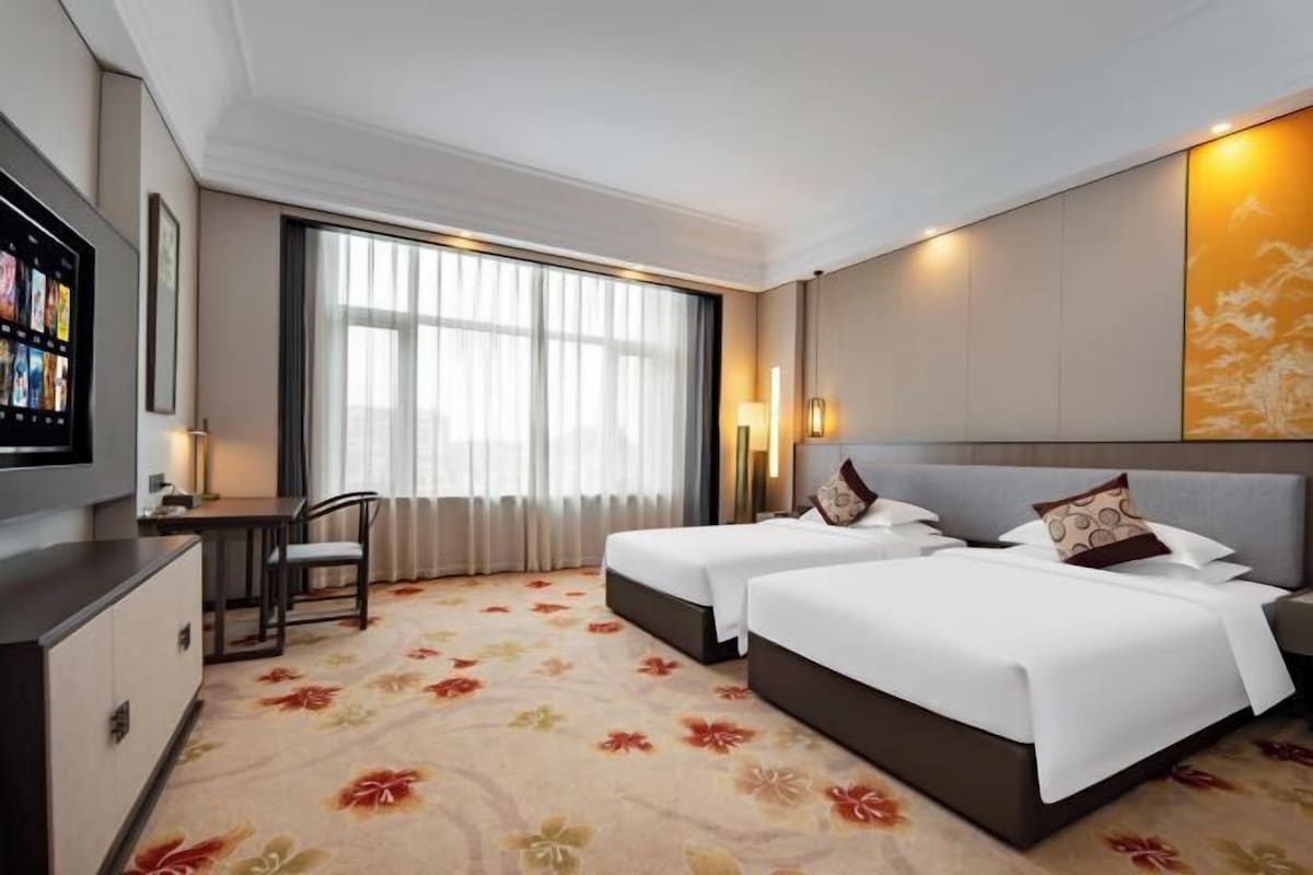 Habitación de hotel con dos camas, un escritorio, un televisor y un ventanal grande con cortinas.