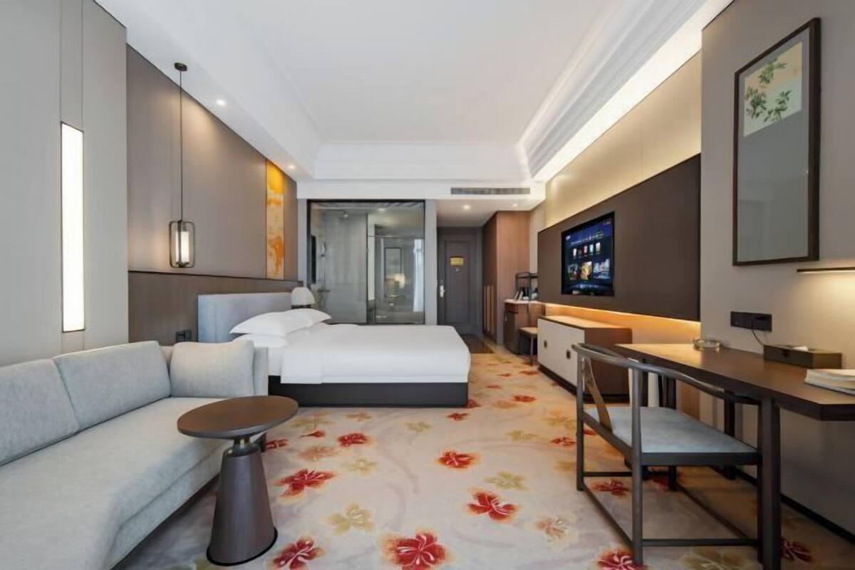 A hotel room with a bed, a sofa, a desk, a TV, and a bathroom visible through a glass door.