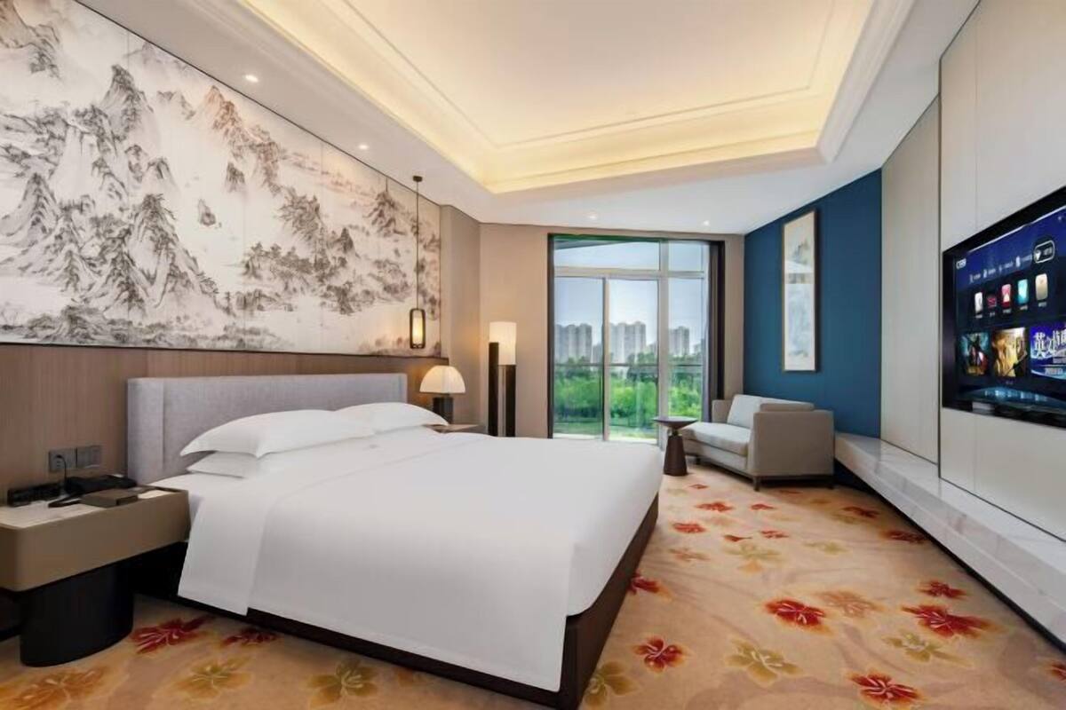 A hotel room with a bed, a desk, a chair, a TV, and a large window.