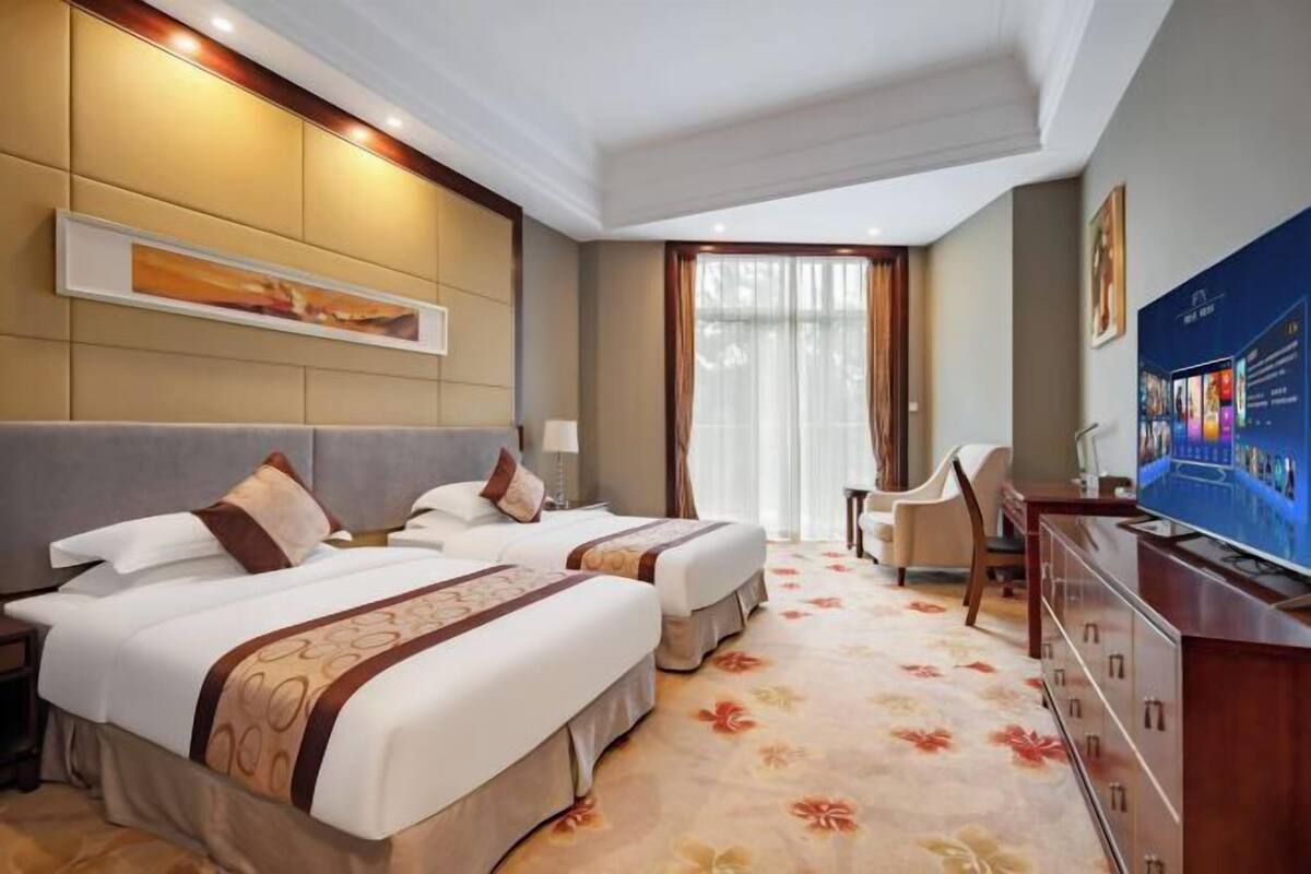 A hotel room with two beds, a desk, a television, and a large window with curtains.