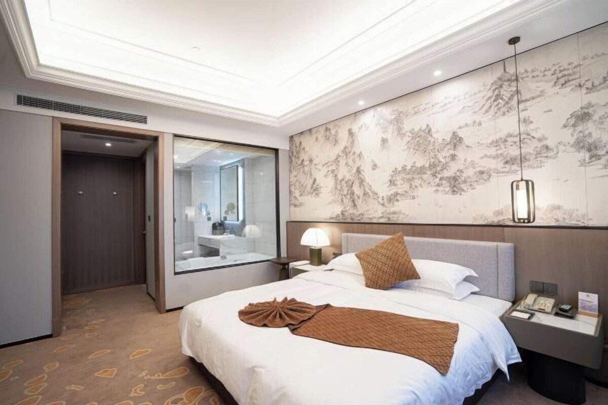 A hotel room with a large bed, a nightstand with a lamp, a wall with a mural, and a door leading to another room.