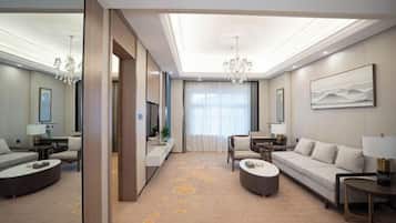 Una habitación de hotel moderna con un sofá grande, otro sofá más pequeño, una mesa de centro y una televisión. La habitación tiene un ventanal con cortinas, un candelabro y un cuadro en la pared.