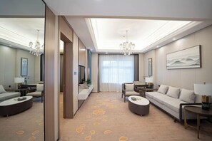 A modern hotel room with a large sofa, a smaller sofa, a coffee table, and a TV. The room has a large window with curtains, a chandelier, and a painting on the wall.