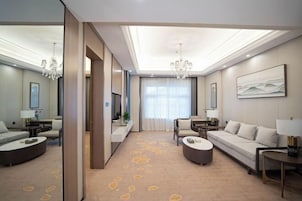 Una habitación de hotel moderna con un sofá grande, otro sofá más pequeño, una mesa de centro y una televisión. La habitación tiene un ventanal con cortinas, un candelabro y un cuadro en la pared.