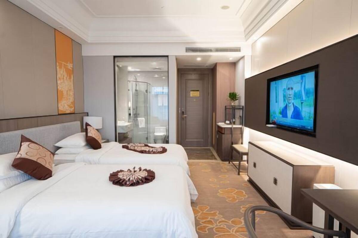 A hotel room with two beds, a TV, a desk, and a bathroom visible through a glass door.