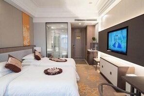 Habitación de hotel con dos camas, televisión, escritorio y baño visible a través de una puerta de vidrio.