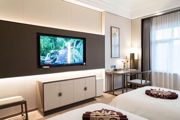 A hotel room with a flat-screen TV mounted on the wall, a desk with a chair, a bed, and a large window with curtains.