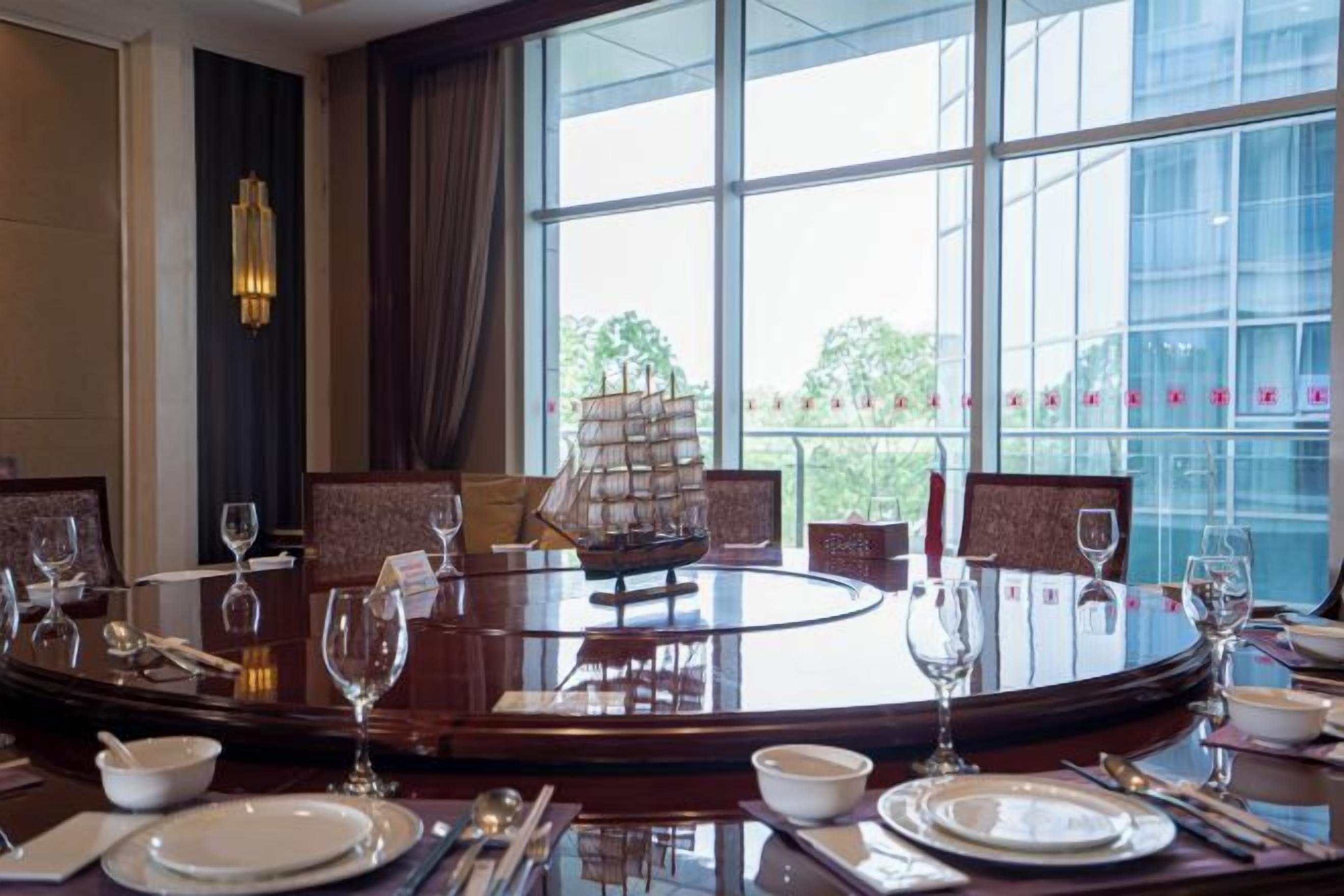 A round dining table with a ship model centerpiece, surrounded by chairs, in a room with large windows and a view of greenery.
