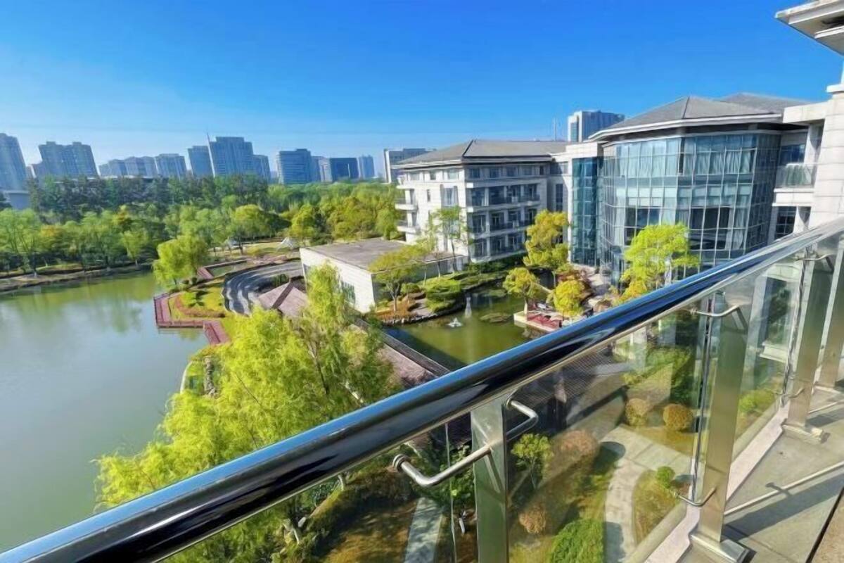 A modern building with a glass facade, a river, and a park with trees and a fountain.