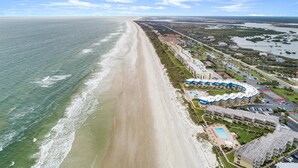 Luftaufnahme eines Küstenhotels mit langer Strandpromenade, Schwimmbad und einem Jachthafen.