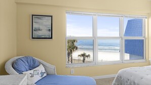 Ein Zimmer mit großem Fenster mit Blick auf einen Strand, ein Korbstuhl mit Kissen und ein gerahmtes Bild an der Wand.