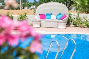 Ein Sitzbereich am Pool mit einer weißen gepolsterten Couch und zwei Beistelltischen, mit Blick auf ein Schwimmbecken.