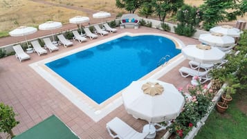 Una piscina rectangular con sillones de playa y sombrillas, rodeada de un patio de ladrillo y follaje.