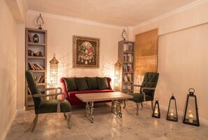 Un saló tranquil i acollidor amb un sofà vermell, dos butaques verdes, una taula de centre de fusta i prestatges plens de llibres i objectes decoratius.