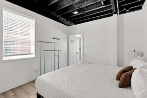A bedroom with a large bed, a window with blinds, a door, and a ceiling with exposed beams.