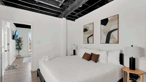 Een moderne slaapkamer met een groot bed, twee abstracte schilderijen, een nachtkastje met een lamp en een televisie in de aangrenzende kamer.