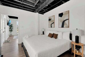 A modern bedroom with a large bed, two abstract paintings, a bedside table with a lamp, and a television in the adjacent room.