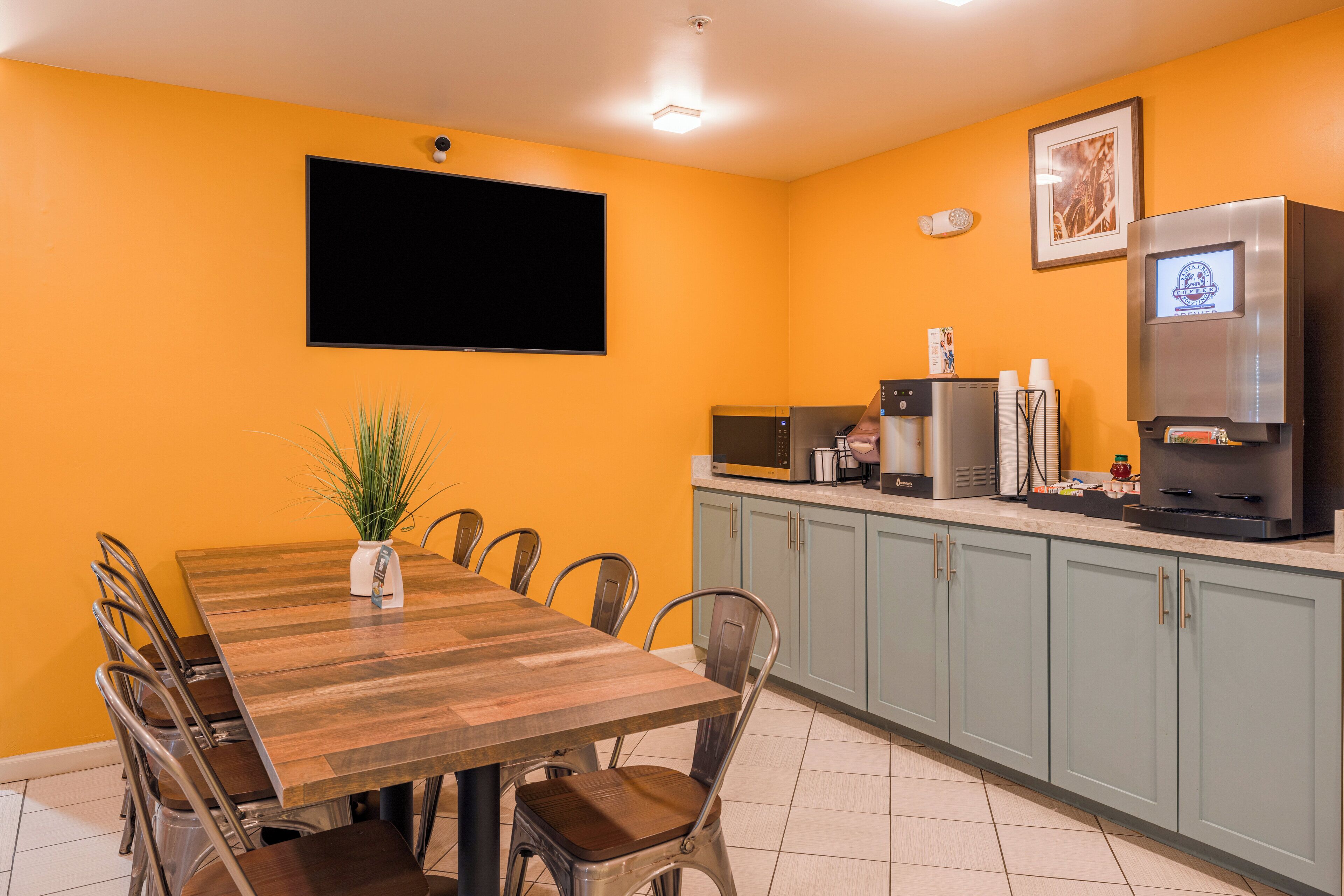 Un comedor con una mesa y sillas de madera, un televisor de pantalla plana montado en la pared y una cocina con microondas y cafetera.