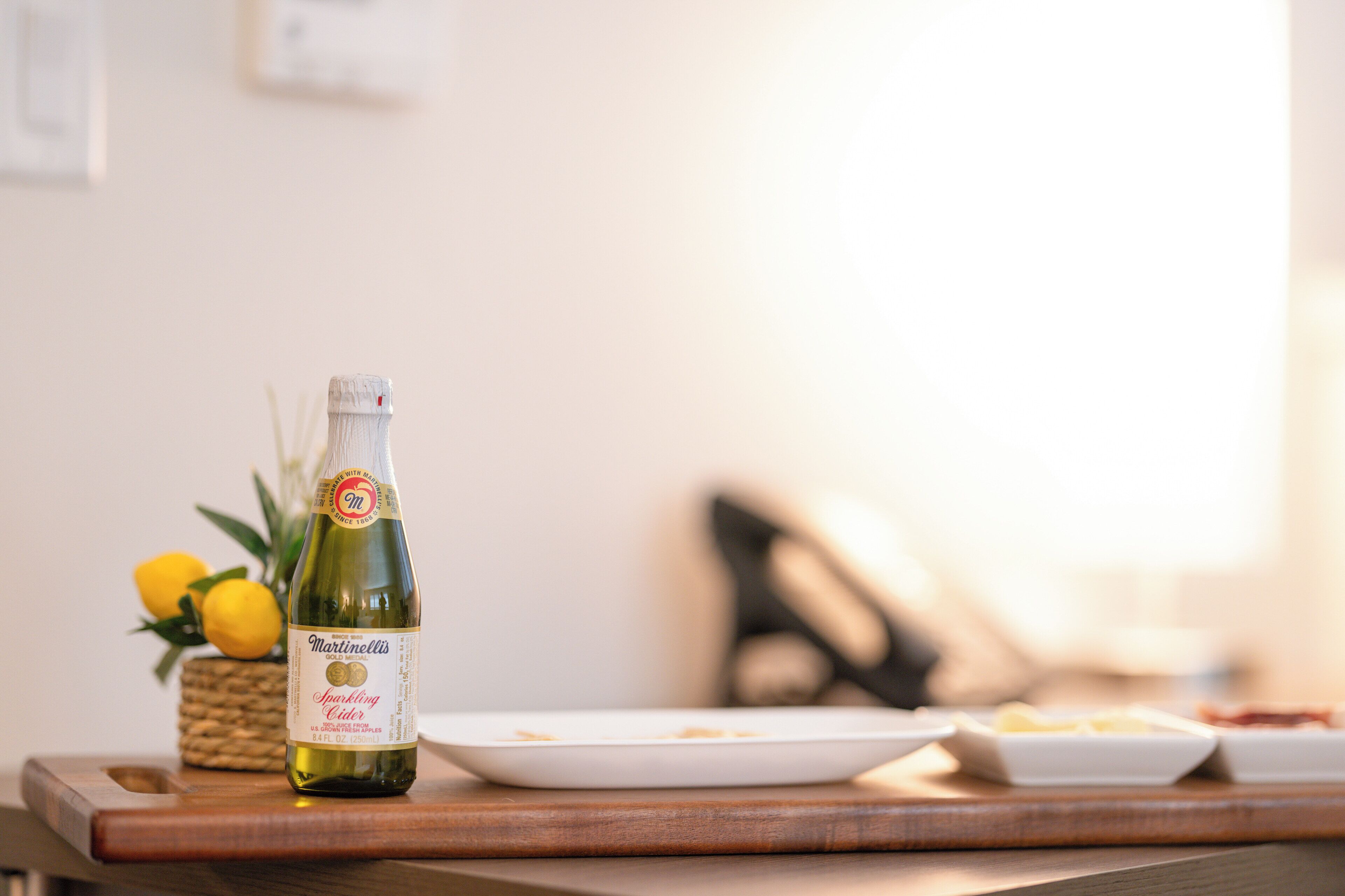 Una botella de sidra espumosa Martinelli sobre una mesa de madera, con un plato blanco y una cesta de limones.