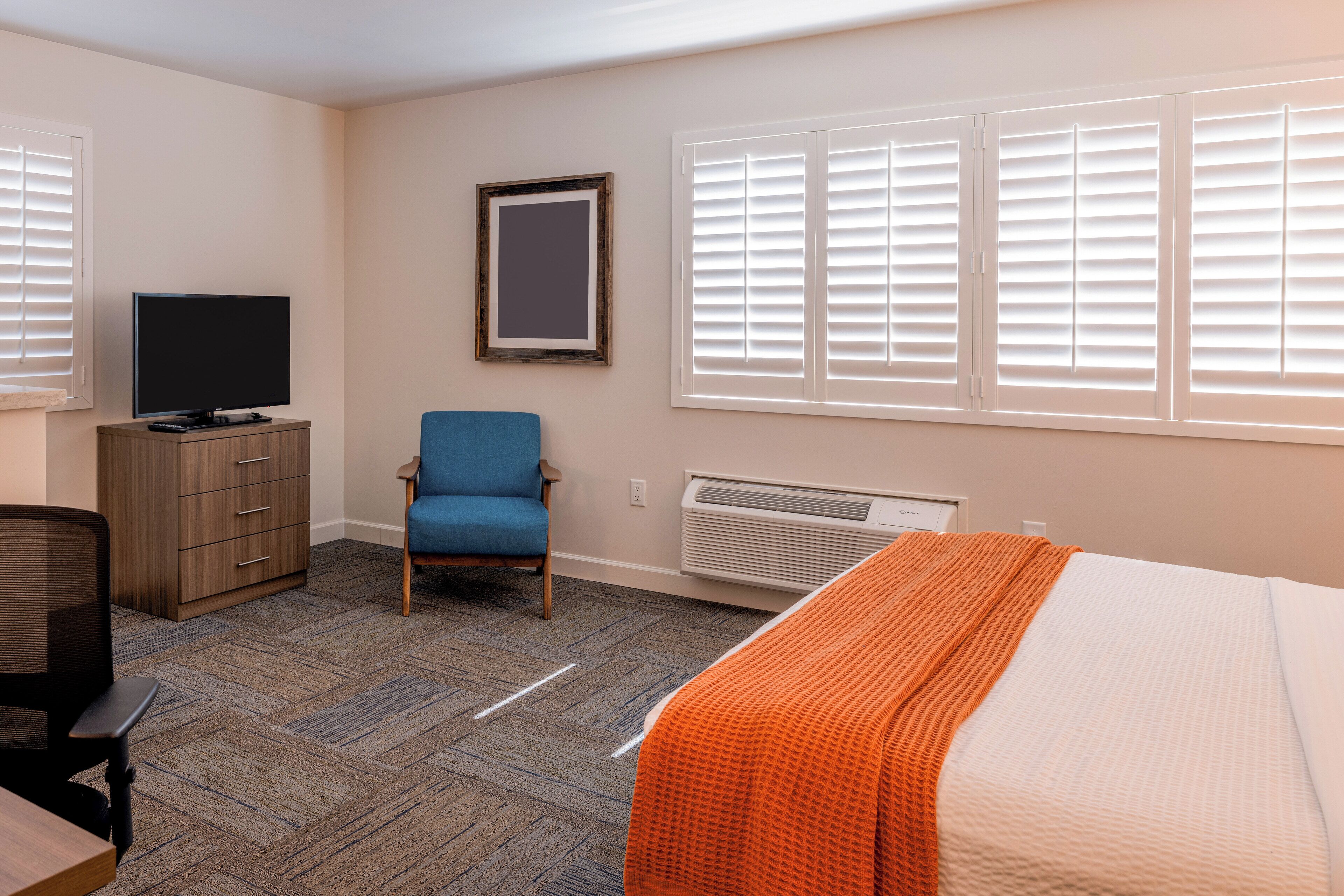 A hotel room with a bed, a desk with a chair, a television, a wardrobe, and a window with blinds.