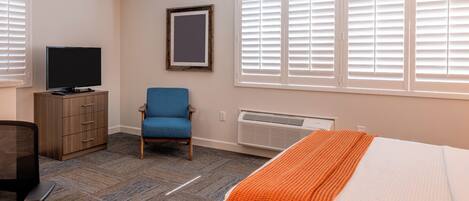 A hotel room with a bed, a desk with a chair, a television, a wardrobe, and a window with blinds.