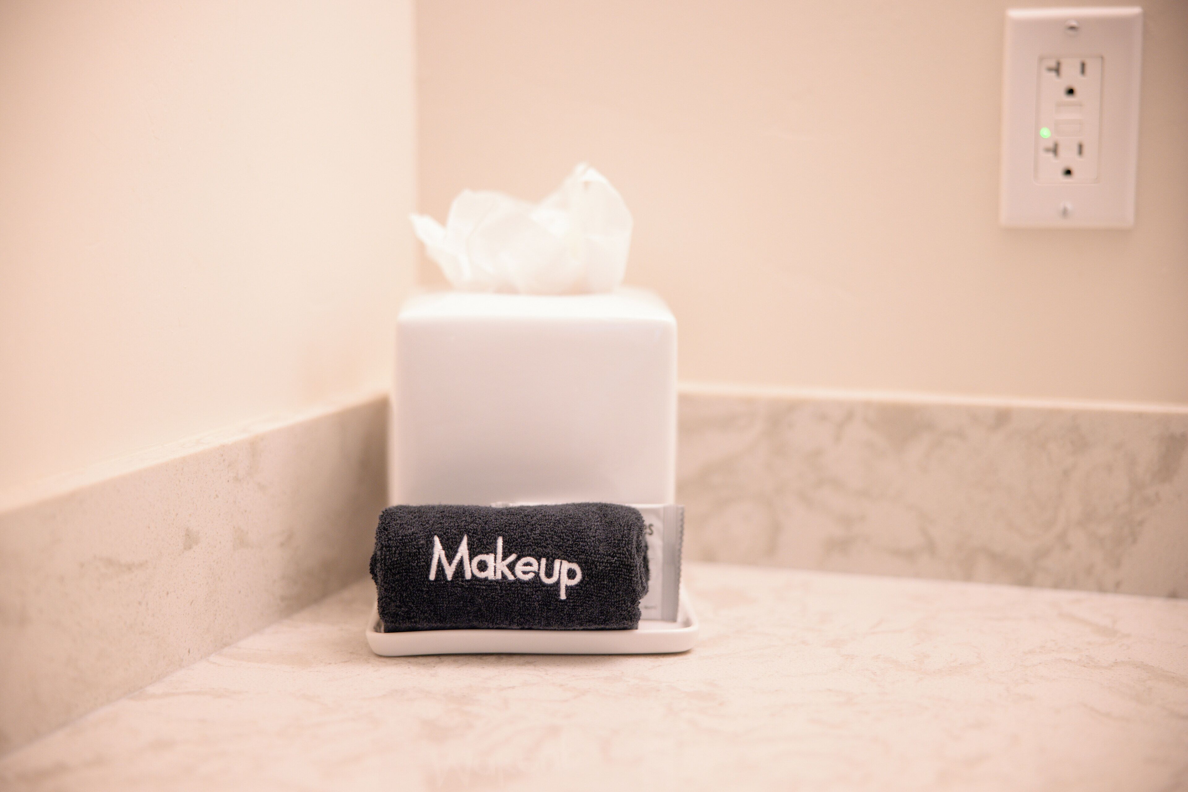 Un coin de salle de bain avec une bo îte de mouchoirs blanche et une serviette noire sur laquelle est inscrit le mot « Maquillage », le tout posé sur un comptoir en marbre.