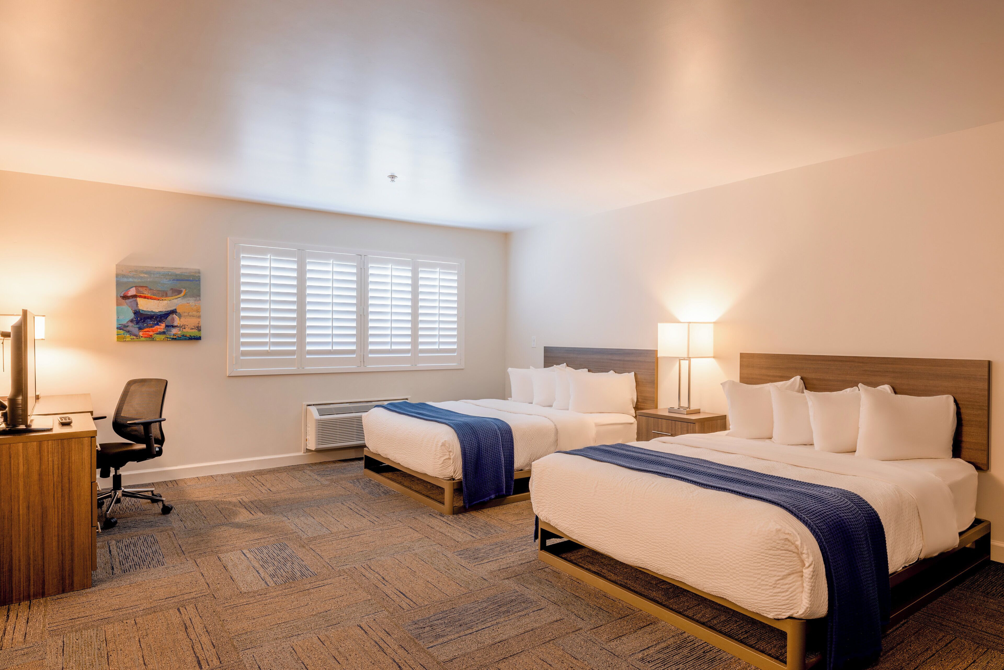 A hotel room with two beds, a desk, a chair, and a painting on the wall.