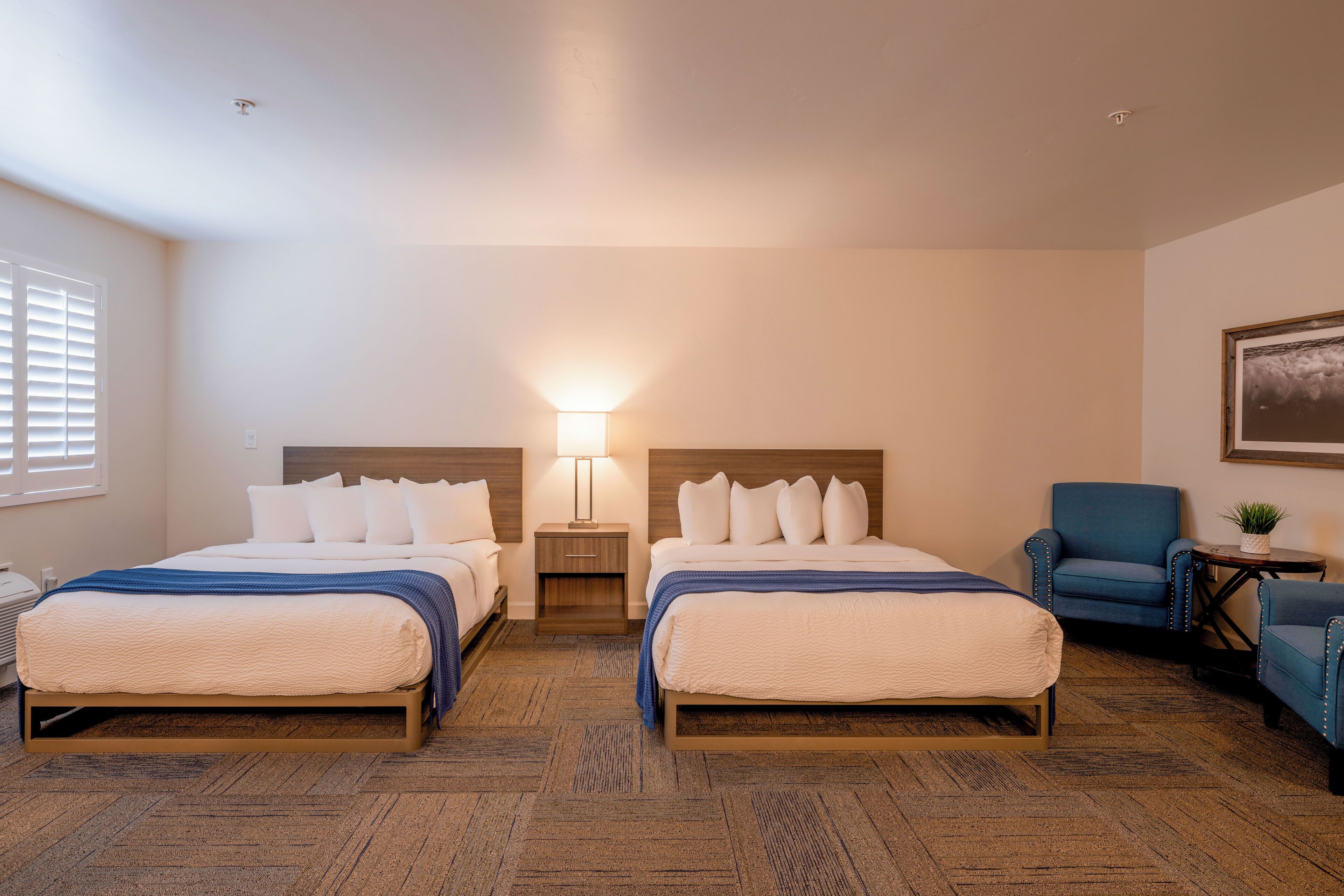 Quarto de hotel com duas camas, uma mesa de cabeceira com abajur, uma cadeira e uma pequena mesa com uma planta.