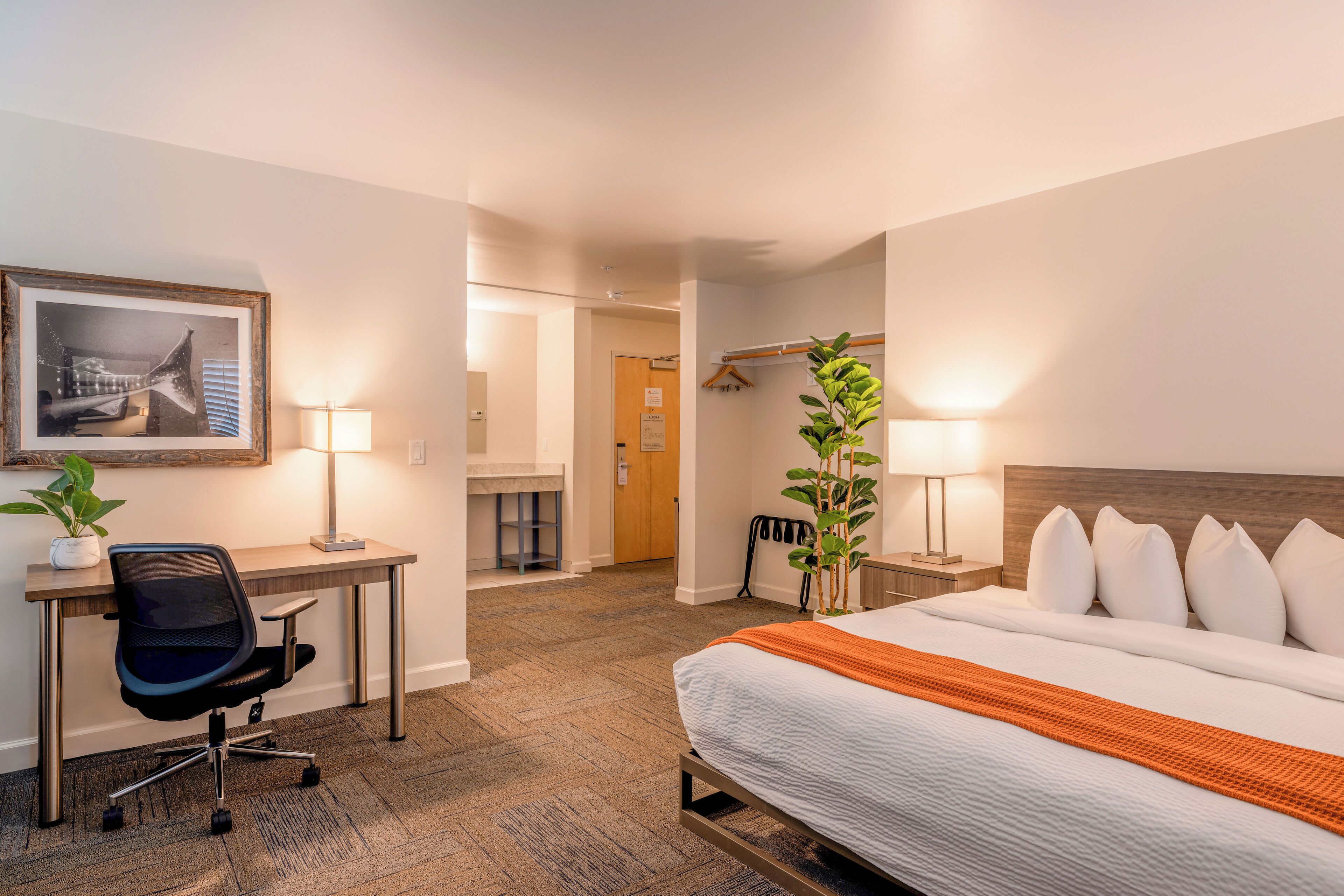 A hotel room with a bed, desk, chair, and a framed picture on the wall.