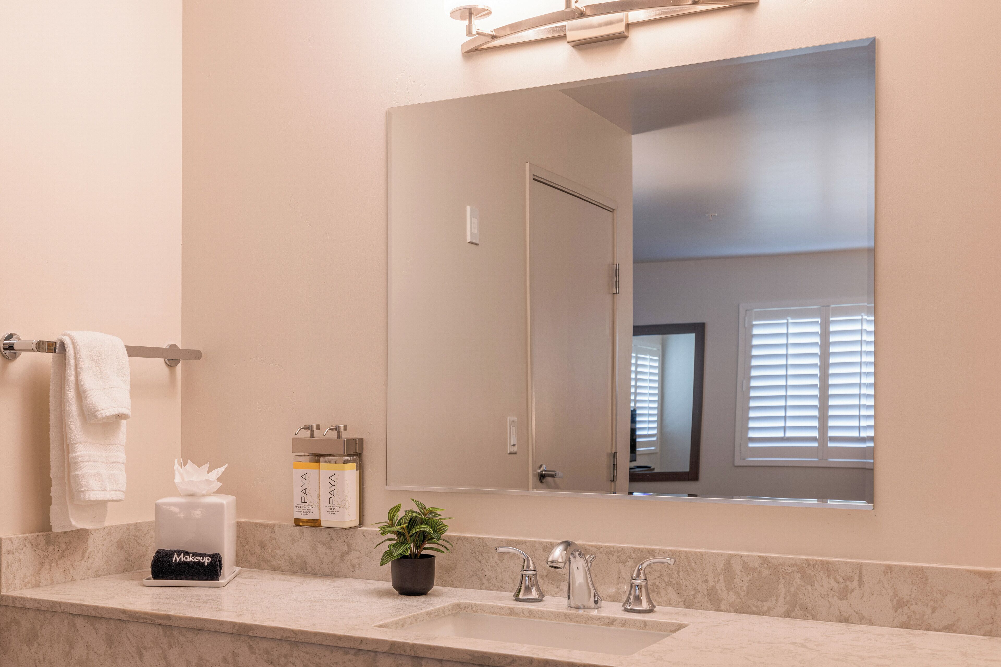 A bathroom with a marble countertop, a sink, a mirror, a towel rack, a potted plant, and a makeup dispenser.