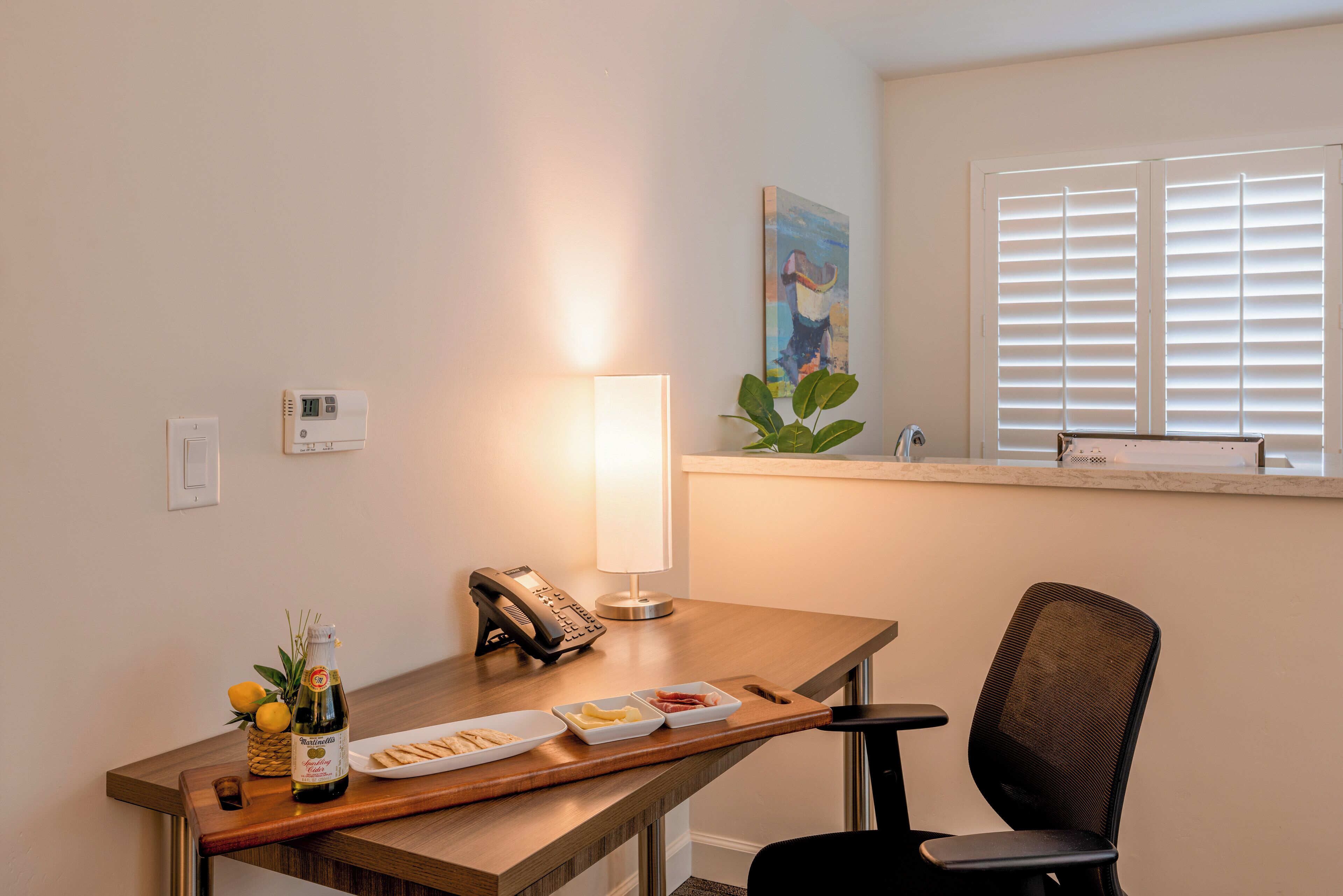 A hotel room with a wooden desk, a lamp, a phone, a small plant, and a window with blinds.