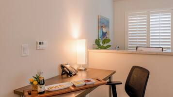 A hotel room with a wooden desk, a lamp, a phone, a small plant, and a window with blinds.