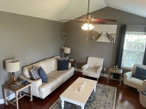 Sala de estar com sofá, poltronas, mesa de centro, abajur, mesa lateral, luminária pendente, ventilador de teto e janela com persianas.