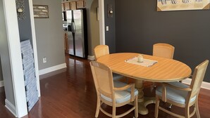 A dining area with a round wooden table and four wicker chairs, a painting of beach chairs on the wall, and a hallway leading to a kitchen with wooden cabinets and a refrigerator. - Wilmington in the middle of it ALL (Wilmington)