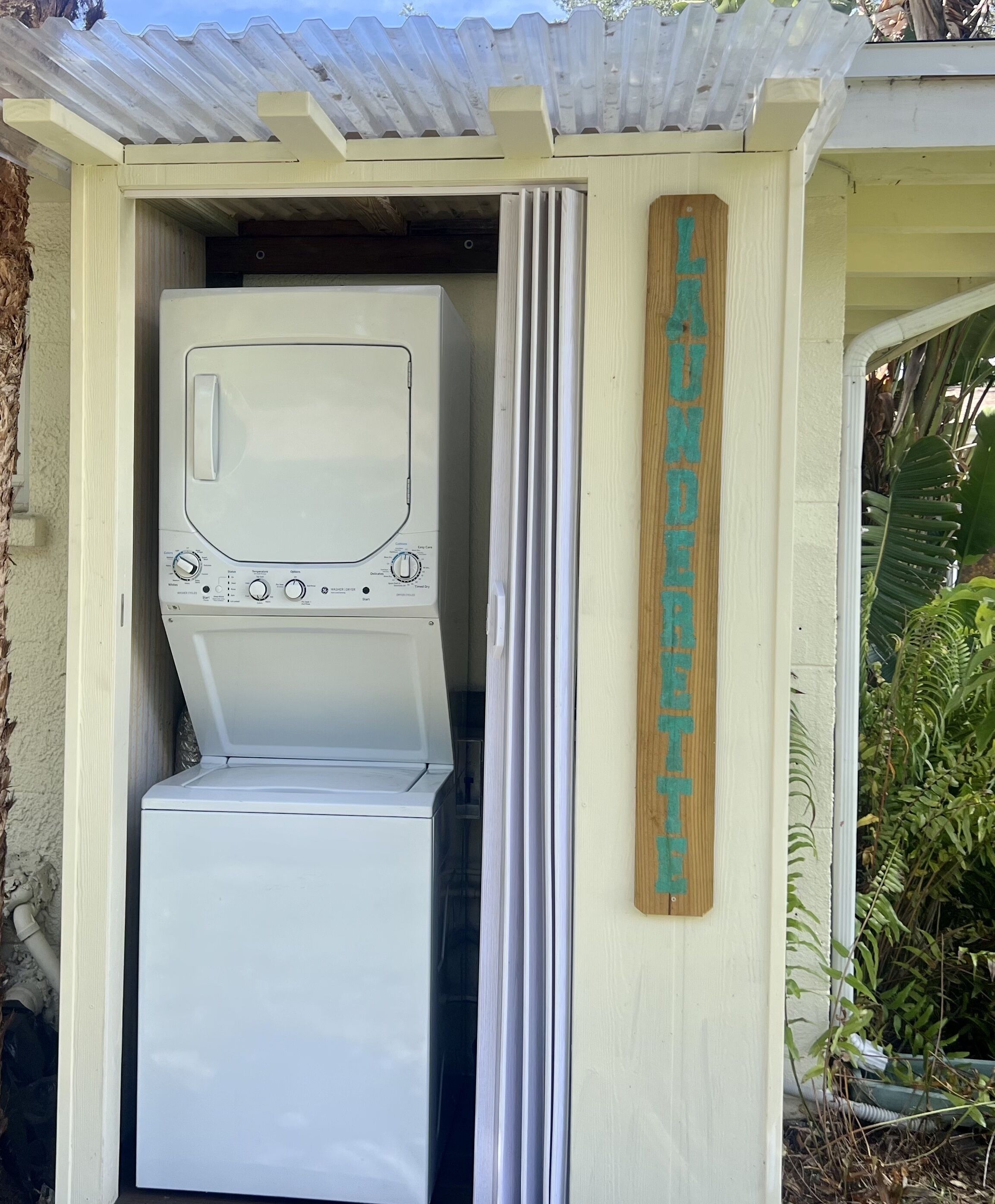 Une buanderie équipée d’un lave-linge et d’un sèche-linge, une enseigne en bois indiquant « Buanderie », et un toit en tôle ondulée.
