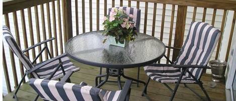 Un patio cubierto con una mesa redonda de vidrio, cuatro sillas a rayas y una planta en maceta.