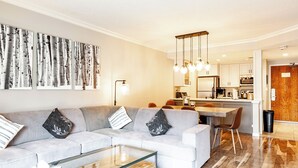 A modern living room with a grey sectional sofa, a glass coffee table, and a kitchen in the background. - Shadow Ridge 401 | Walk to Lifts | Mtn & Ski Views | Kitchen | Fireplace | Updated | Pool & Hot Tub (Park City)