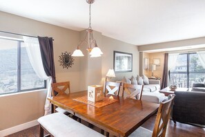 A dining area with a wooden table, chairs, and a chandelier. There is a window with curtains and a view of the outside.