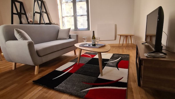 Una sala moderna con un sofá gris, una mesa de centro con una botella de vino, un televisor sobre un mueble de madera y un ventanal con vistas.