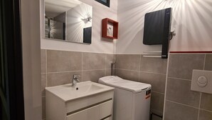 Un baño moderno con un lavamanos blanco, un espejo y un inodoro. Hay una lámpara en el techo y un cuadro pequeño con marco rojo en la pared.