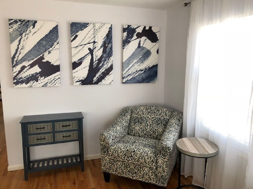 A corner room with a patterned armchair, a small side table, and three abstract paintings on the wall.