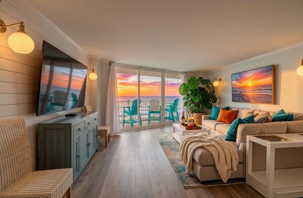 A living room with a TV, sofa, and chairs, a balcony with a view of the sunset, and a dining area with a table and chairs.