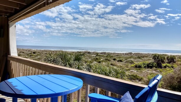 A balcony with two blue chairs and a small table, overlooking a beach with sparse vegetation and a clear sky.