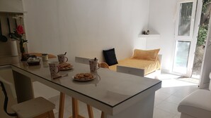 Une cuisine moderne avec un îlot central en verre, des tabourets en bois et une table à manger. On y trouve un lit avec une tête de lit jaune et un canapé blanc dans l’espace salon.