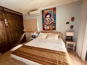 A hotel room with a large bed, a wooden wardrobe, a nightstand with a lamp, and a colorful artwork on the wall.